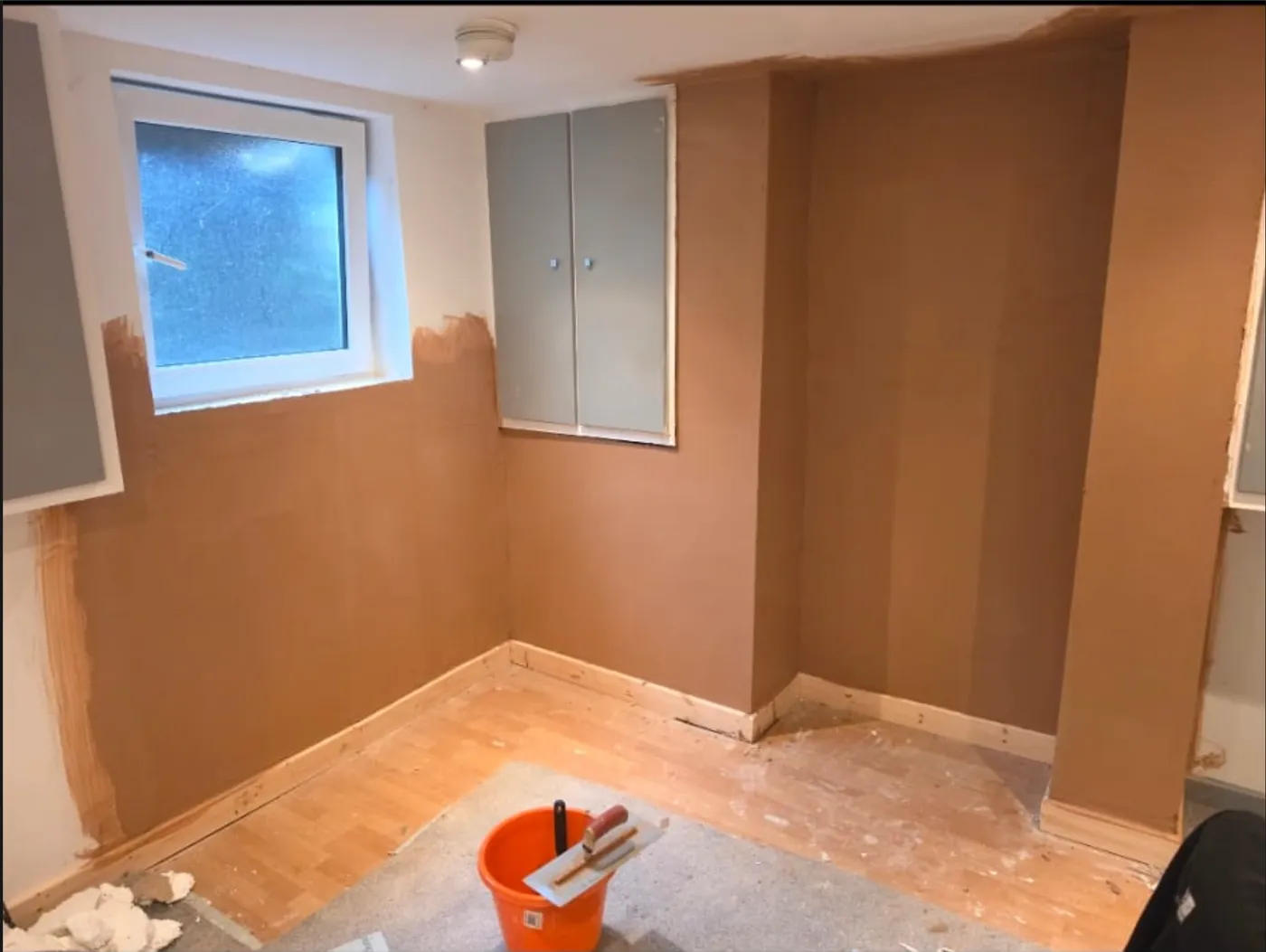 A room replastered throughout, the new render still drying, with a trowel and bucket left on the boards.
